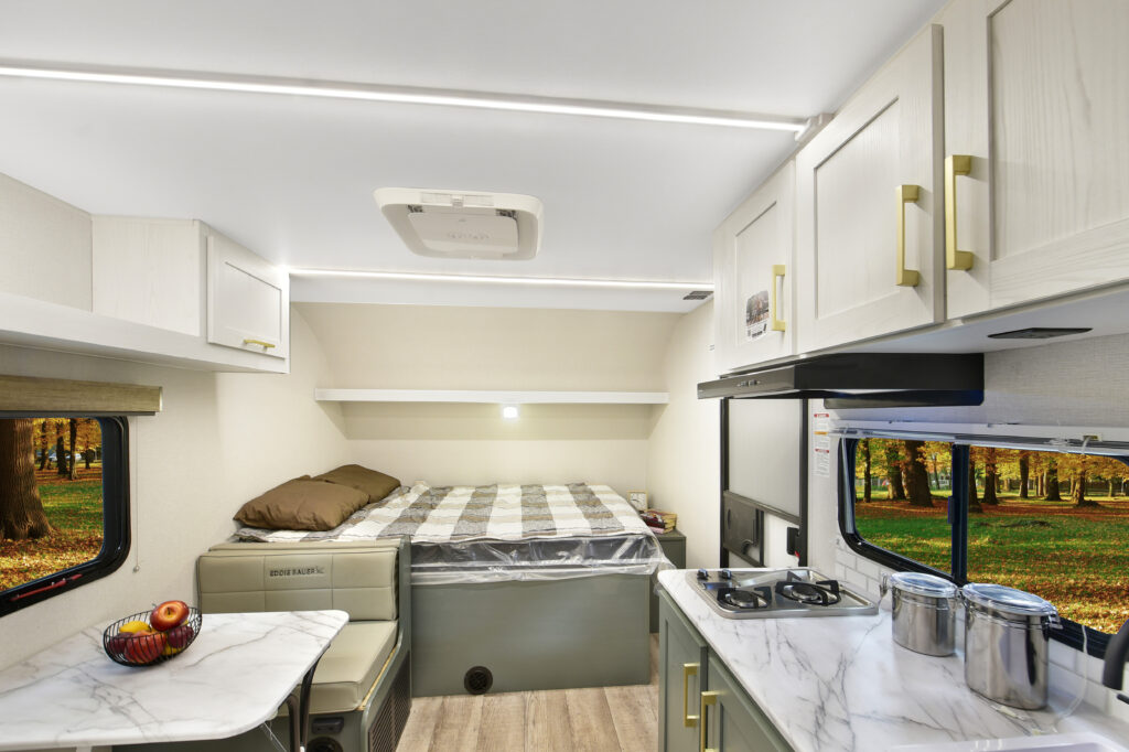 Interior of a camper van featuring a bed with plaid bedding, a small kitchen with a stove and counters, cabinets, and windows showing a view of trees and grass outside. A bowl with apples sits on the counter.