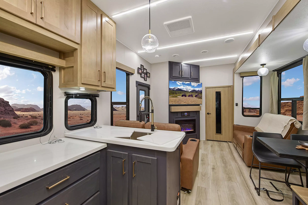 Experience the modern 2025 RV interior designed for families, featuring a kitchen with light wood cabinets, white countertops, a sink, and a stove. The living area boasts a sofa and mounted TV. Large windows reveal scenic desert landscapes with rock formations while ceiling lights illuminate the space.