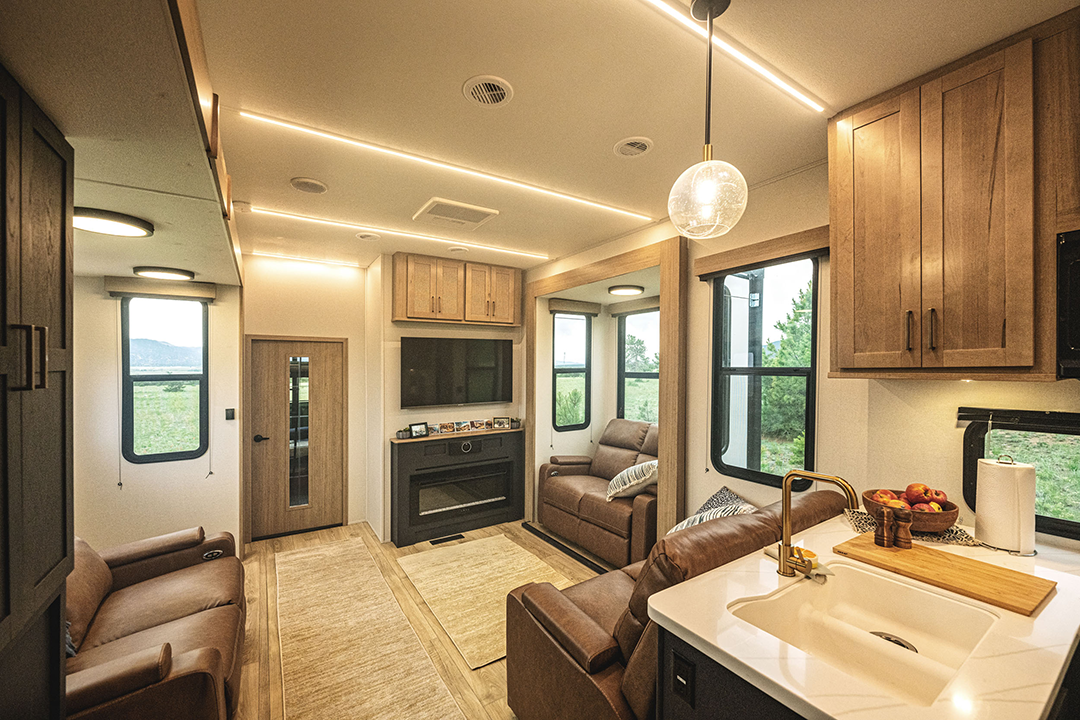The interior of this 2025 RV toy hauler boasts a cozy lounge with two sofas, a fireplace beneath a mounted TV, and large windows. A kitchen counter with a sink and fruit basket sits upfront. Bright lighting paired with warm wooden tones crafts a welcoming ambiance.