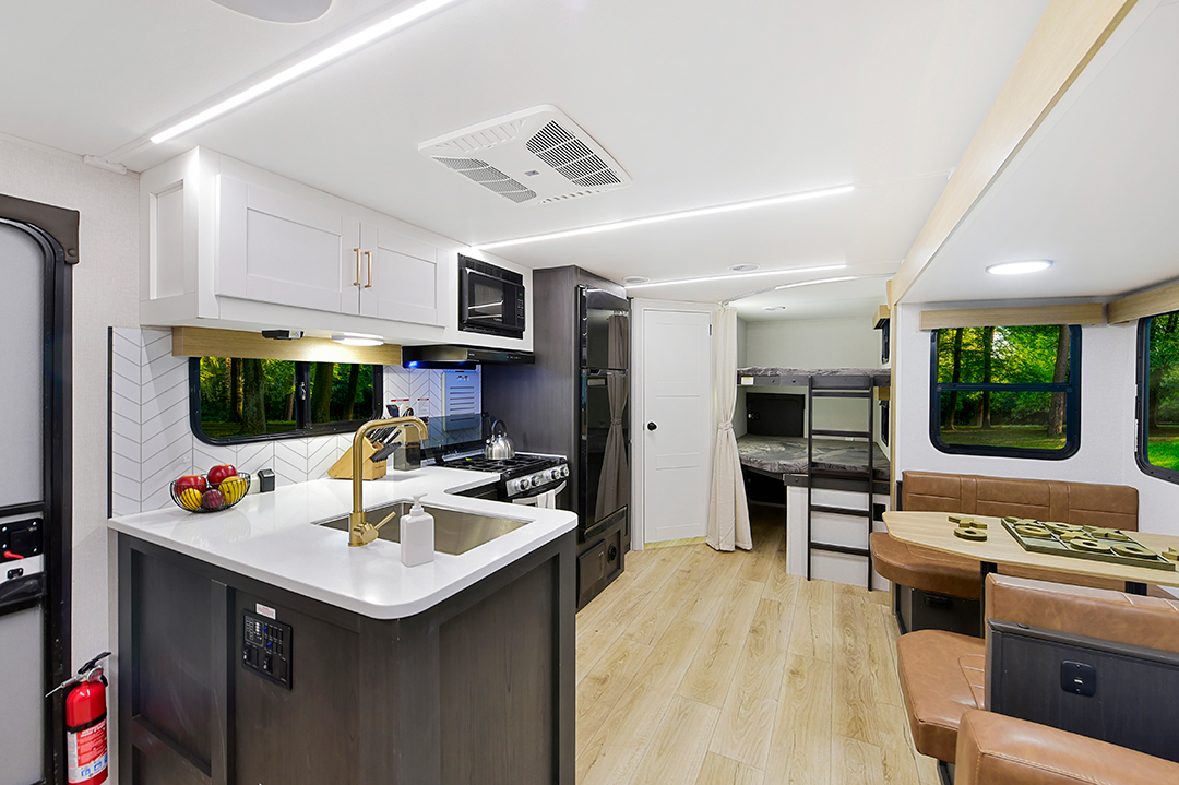 In the 2025 family-friendly RV interior, youll find a modern kitchen with a sleek sink, stove, microwave, and white cabinets. To the right is a cozy dining area with cushioned seating, while bunk beds lie in the background perfect for families. Large windows reveal a lush green landscape beyond.