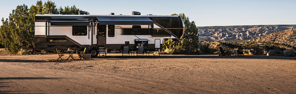 A large modern Bighorn RV is parked on a dirt clearing near trees, with outdoor chairs and a table set up. In the background, there are hills and a wide, open landscape under a clear sky.