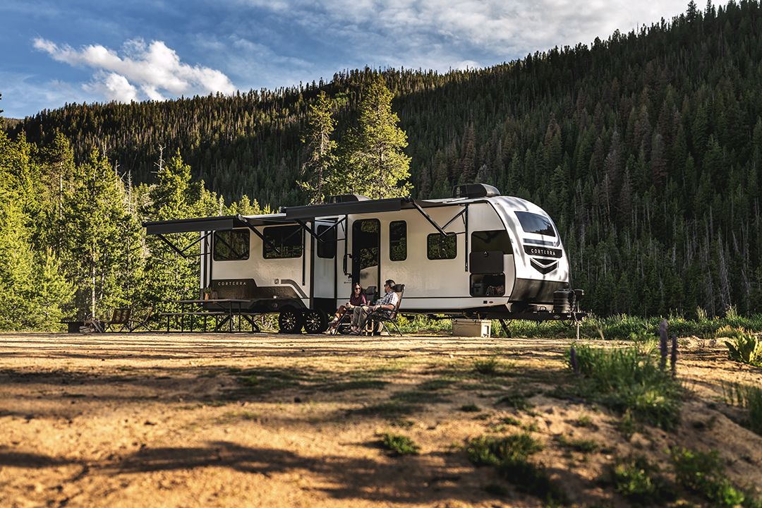 A large RV with auto-leveling is parked in a scenic forest clearing. Two people sit in camping chairs under the RV’s awning, surrounded by tall pine trees and mountains as sunlight highlights the peaceful, natural setting.