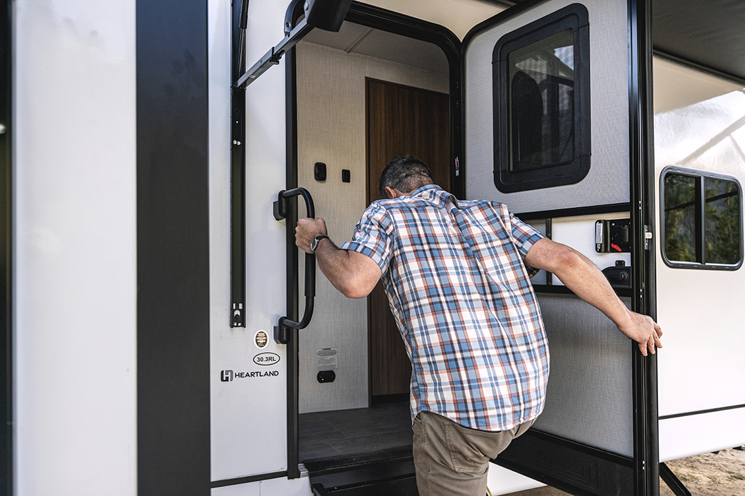 A man wearing a plaid shirt and khaki pants is holding onto a handle while stepping into a white Heartland RV with auto-leveling through its open door.
