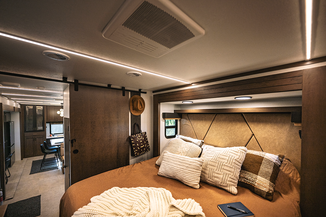 Interior of a cozy Bighorn RV bedroom with a neatly made bed, multiple pillows, and a blanket. A sliding wooden door separates the bedroom from the dining space in the main living area. Natural light filters through small windows.