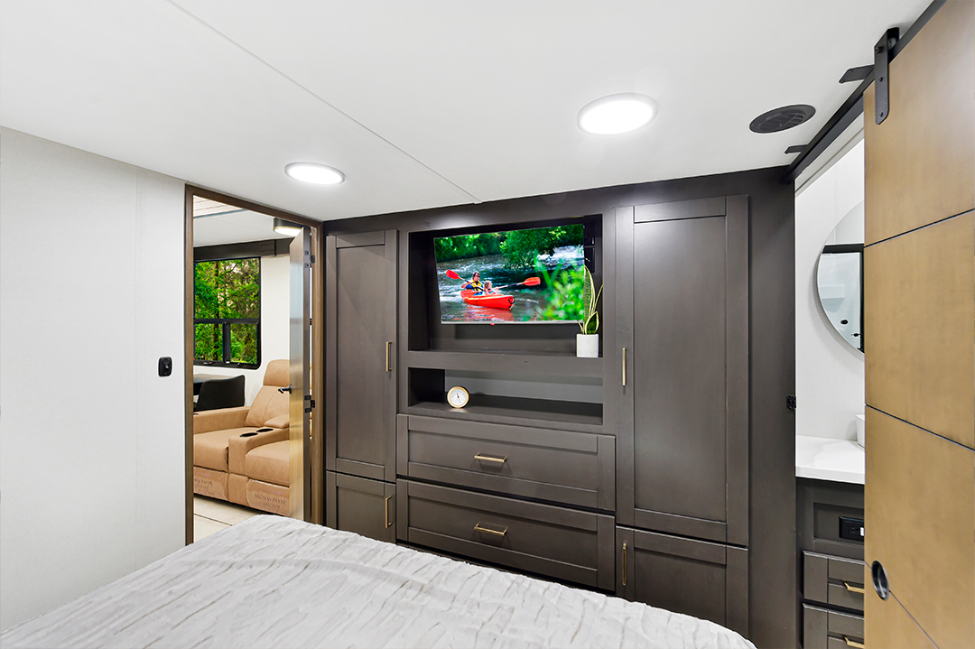 Modern RV bedroom with a bed in the foreground, built-in dark wood cabinetry, and a TV displaying people kayaking—offering comfort and style often found in popular bunkhouses. A beige sofa and green trees are visible through an open door.