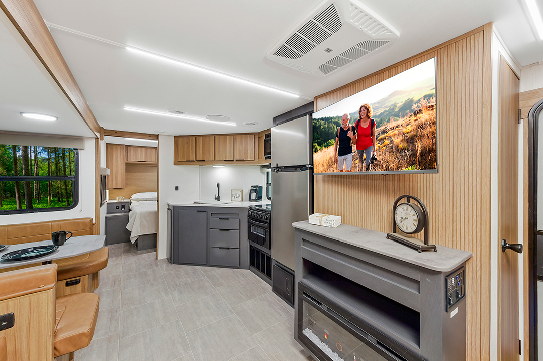 Innovative travel trailer interior with kitchen, dining area, and bed. A large flat-screen TV displays two people outdoors. Stylish wood accents, gray cabinets, and a fireplace with decor complete the space. Large window reveals green trees outside.