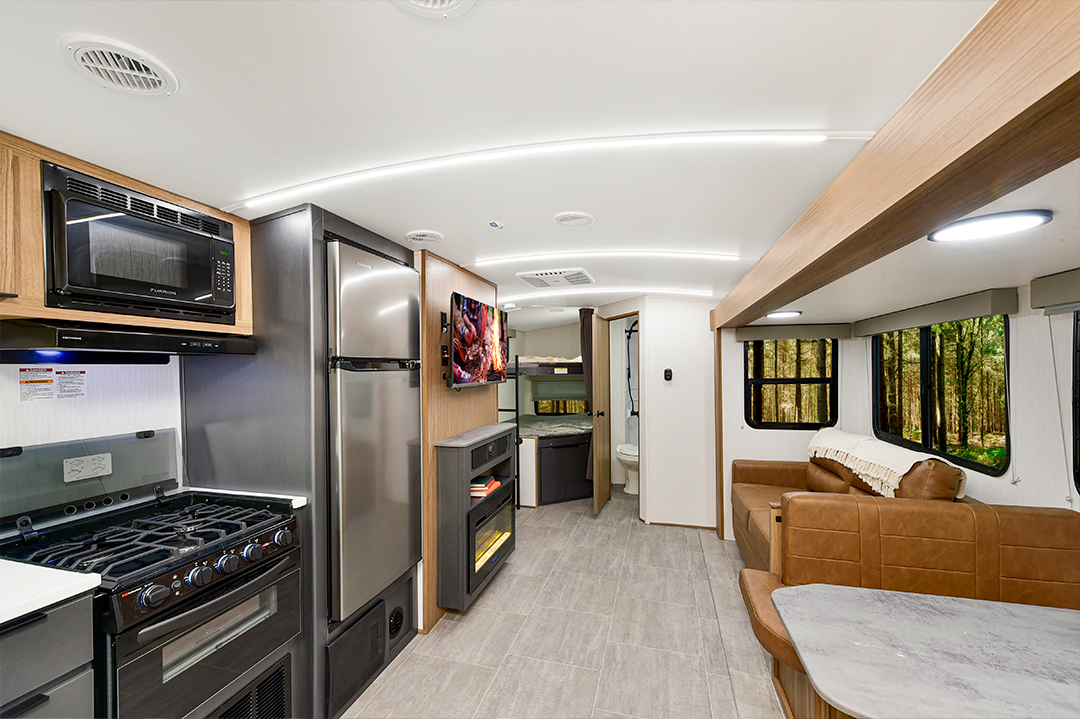 Modern RV interior featuring a kitchen with a stove, oven, microwave, and fridge, a living area with TV, electric fireplace, brown sofa, and large windows overlooking the forest. Spacious bunkhouses and a bathroom are visible at the rear.