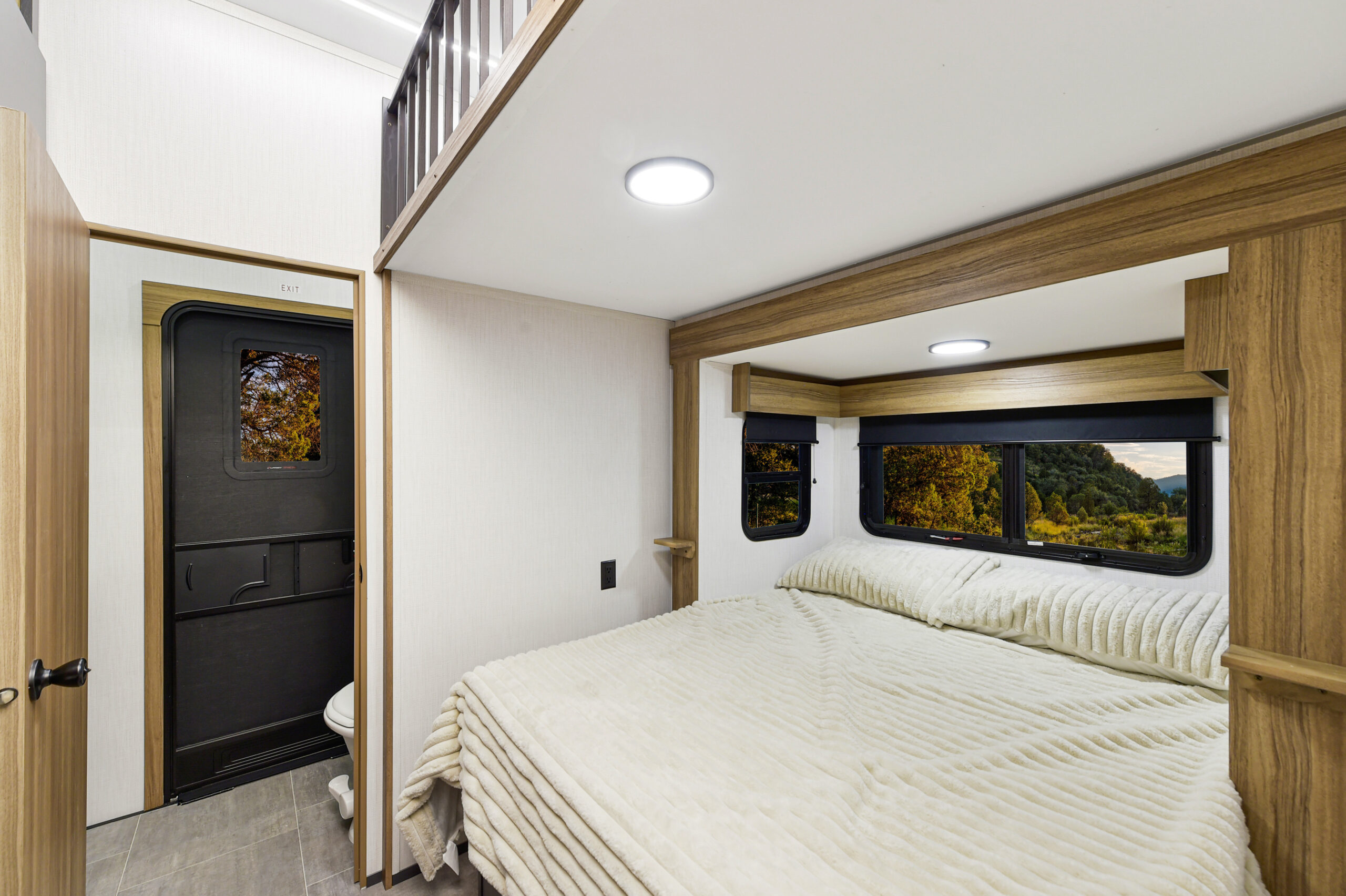 A modern RV bedroom with a large bed, light-colored bedding, wood accents, and windows revealing a scenic forest view. On the left, a small bathroom with a closed black door is visible—perfect for families exploring bunkhouses or spacious RV layouts.