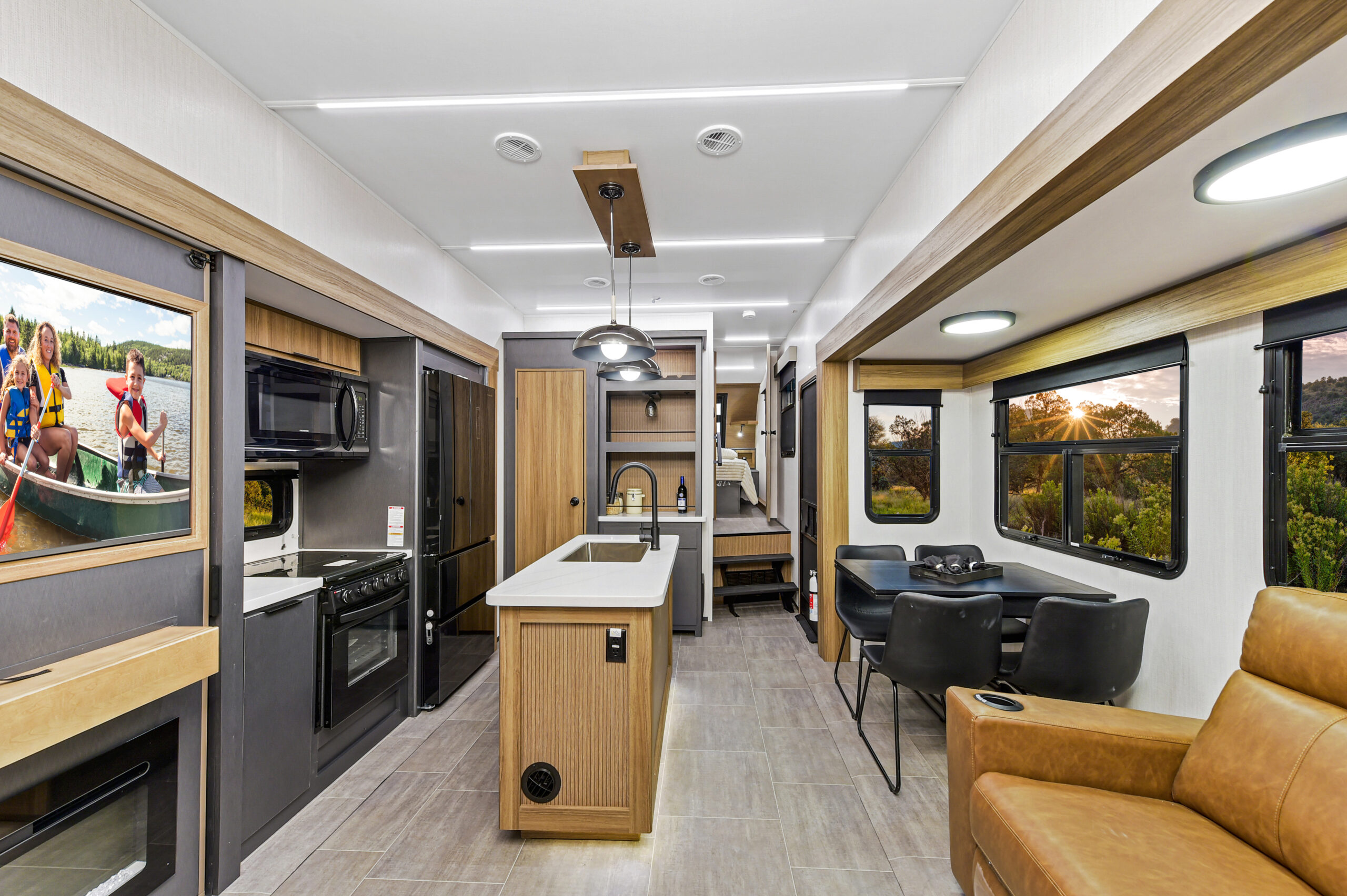 Modern RV interior with a kitchen, dining table, leather chair, and large windows showing a sunset view. A TV displays people canoeing. The bright decor features wood accents, stainless steel appliances, and spacious bunkhouses for added comfort.