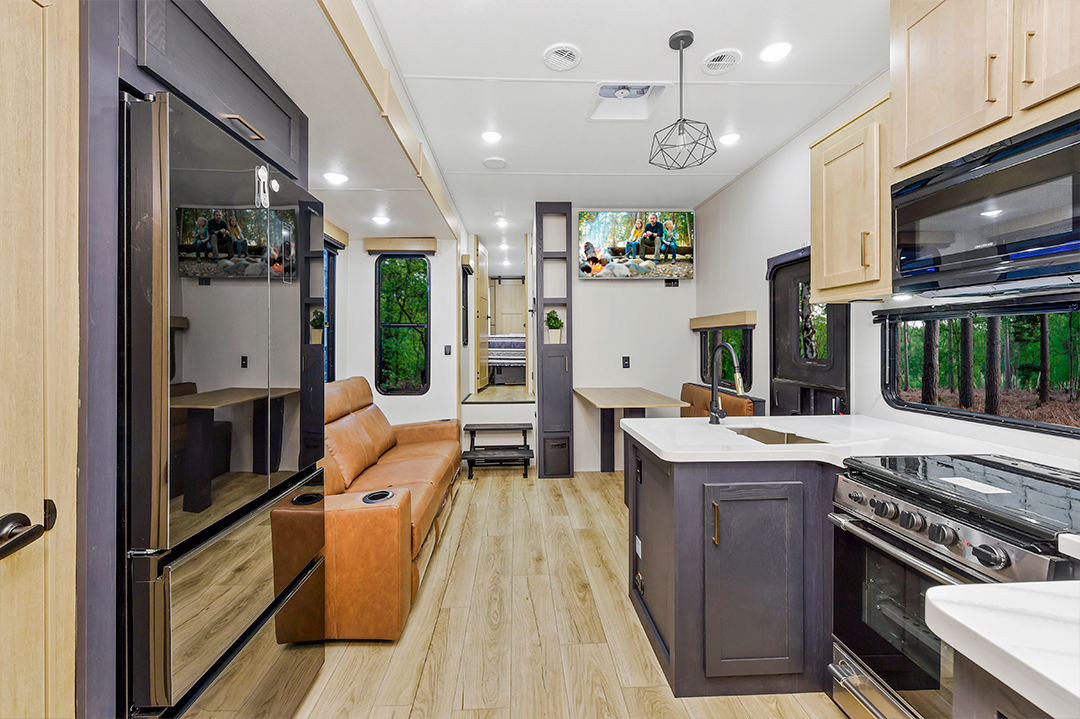 Modern RV interior with light wood floors, a brown leather sofa, a kitchen area with stove, sink, and refrigerator, a dining table, and large windows showing green trees. Bunkhouses offer extra sleeping space. A TV is mounted above the sofa.