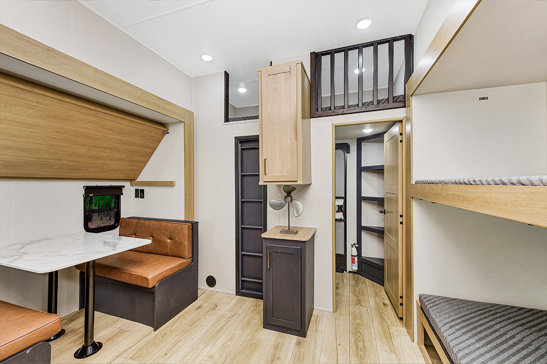 Modern RV interior with light wood floors, booth dinette with brown cushions, compact kitchen, and cozy bunkhouses on the right. A loft above the central door adds extra space to this clean and organized design.