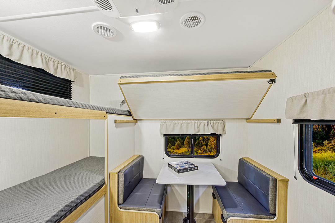 Interior of a camper van showing a dining area with a table and four seats, two bunkhouses-style bunk beds, and a window with a nature view. A book and glasses rest on the modern, minimal table décor.