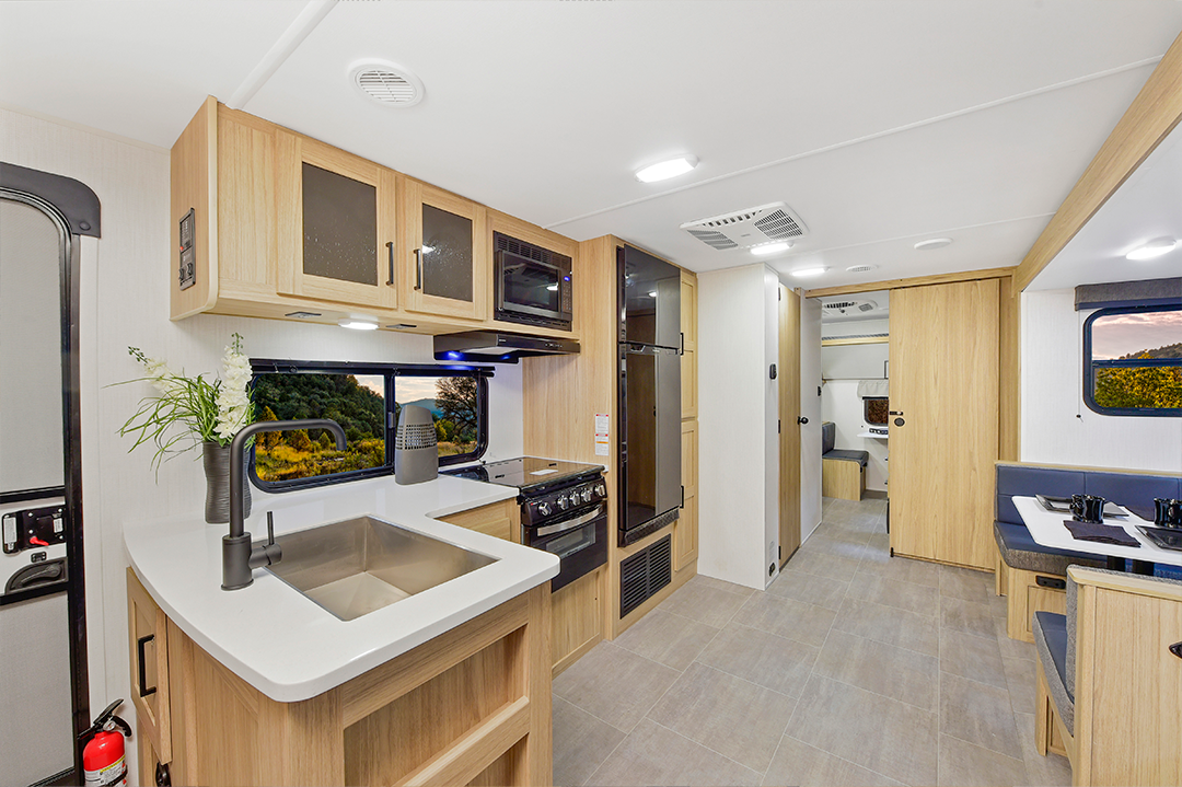 Modern RV interior with light wood cabinets, a kitchen area featuring a sink, stove, and microwave, a dining booth on the right, and a hallway leading to bunkhouses, bedroom, and bathroom. Large windows reveal scenic outdoor views.