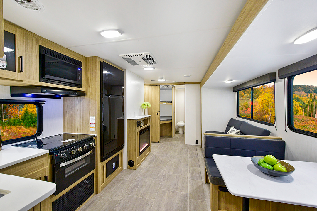 Modern RV interior with wood accents, featuring a kitchen with stove and microwave on the left, a dining area and sofa on the right, and bunkhouses for extra sleeping space. Large windows reveal autumn trees outside and a bathroom at the back.