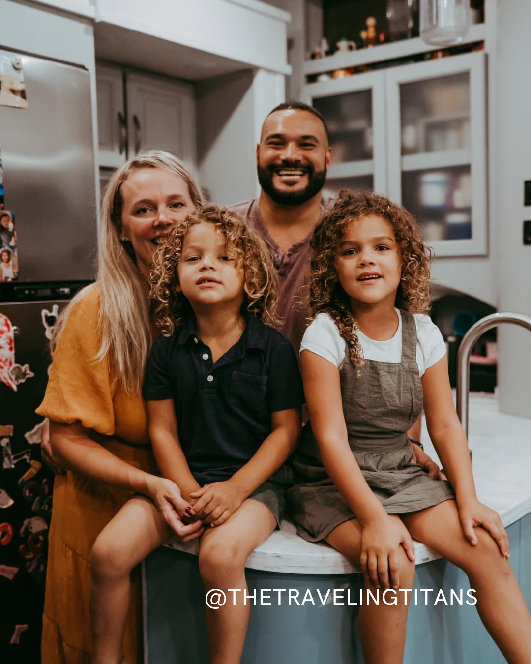 A smiling family of four poses in their kitchen. Two adults stand behind two young children sitting on a counter. The text @THETRAVELINGTITANS is overlaid at the bottom of the image.