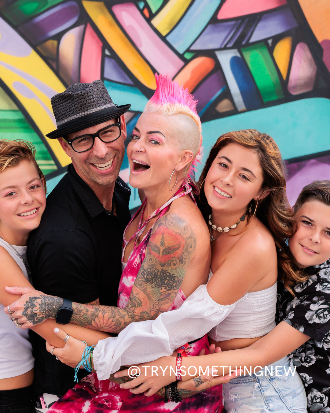 A joyful group of four people, including a woman with a pink mohawk and tattoos, pose together in front of a colorful graffiti wall, smiling and hugging closely. Text at the bottom reads @TRYNSOMETHINGNEW.