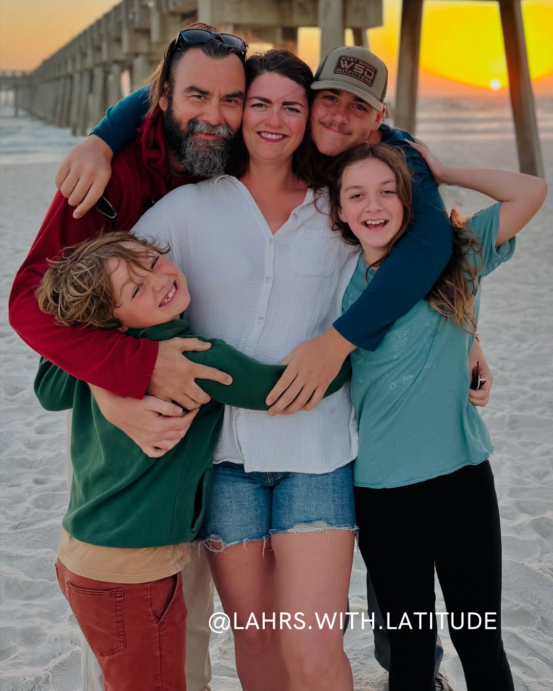 A smiling family of five hugs on a sandy beach at sunset, with a pier extending into the ocean in the background. The group appears happy and relaxed together. Text at the bottom reads @LAHRS.WITH.LATITUDE.