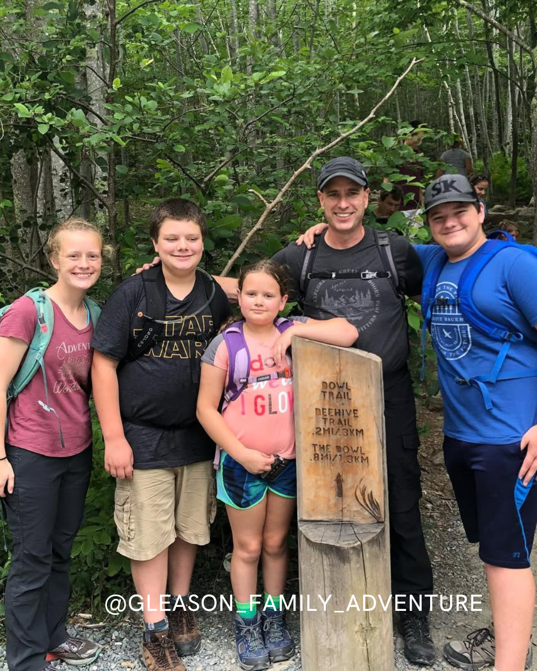 A family of five poses together at a forest trailhead, smiling next to a wooden sign. Trees and greenery surround them, and theyre dressed in hiking clothes with backpacks. Text @GLEASON_FAMILY_ADVENTURE appears at the bottom.